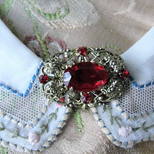 Op de afbeelding: Een vintage broche met een grote rode ovale steen omringd door kleinere rode stenen en ingewikkeld goudkleurig metaalwerk.