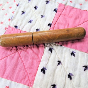 Könnte beinhalten: Ein hölzerner Fingerhut mit natürlicher Oberfläche. Der Fingerhut ist an einem Ende geschlossen und am anderen offen. Er liegt auf einem rosafarbenen und weißen Patchwork-Steppbett.