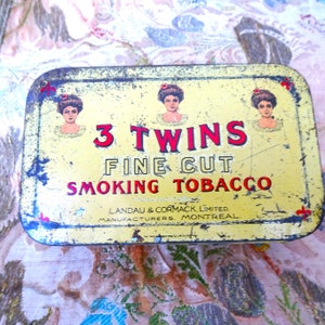 May include: Vintage yellow tin with a red and white label that reads "3 Twins Fine Cut Smoking Tobacco". The label features three images of women with red flowers in their hair.