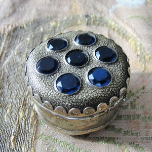 May include: A small, round, silver trinket box with a scalloped edge and a textured surface. The lid is decorated with eight blue gemstones.