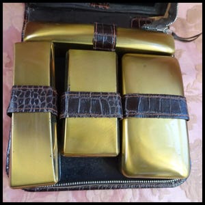 Puede incluir: Un estuche de viaje vintage de cuero con cuatro recipientes de metal de color dorado. Los recipientes tienen correas de cuero marrón con un patrón de cocodrilo.