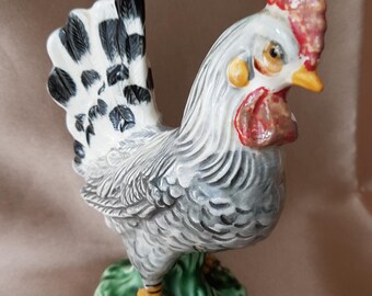 Pode incluir: Uma figura de galo de cerâmica com corpo branco e cinza, penas de cauda pretas e brancas, crista vermelha e pés amarelos. O galo está em pé sobre uma base verde.