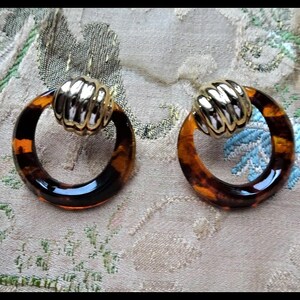 May include: A pair of earrings featuring a circular, brown and black tortoiseshell-patterned hoop attached to a polished, ribbed gold-tone metal stud. The earrings have a high-gloss finish and a classic, geometric design with contrasting textures.