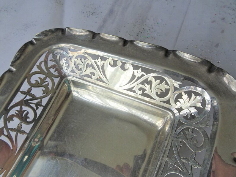 K&ouml;nnte beinhalten: Ein rechteckiges Serviertablett aus Silber mit gewelltem Rand und aufwendigen durchbrochenen floralen und scrollartigen Designs. Das Tablett hat eine hochglanzpolierte, reflektierende Oberfl&auml;che, und der dekorative Rand verleiht dem St&uuml;ck eine elegante Note.