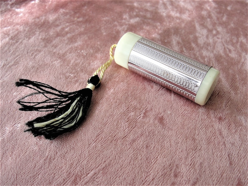 May include: A silver and white cylindrical container with a black and white tassel attached to it. The container has a textured surface and is lying on a pink velvet surface.