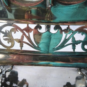 K&ouml;nnte beinhalten: Nahaufnahme eines silberfarbenen Serviertabletts mit aufwendigen, ausgeschnittenen Filigranarbeiten. Die Oberfl&auml;che des Tabletts reflektiert T&uuml;rkis- und Kupfert&ouml;ne. Das Design umfasst florale und schildartige Formen, die einen Vintage- oder Antik-Stil suggerieren.