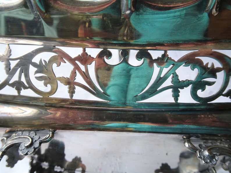 K&ouml;nnte beinhalten: Nahaufnahme eines silberfarbenen Serviertabletts mit aufwendigen, ausgeschnittenen Filigranarbeiten. Die Oberfl&auml;che des Tabletts reflektiert T&uuml;rkis- und Kupfert&ouml;ne. Das Design umfasst florale und schildartige Formen, die einen Vintage- oder Antik-Stil suggerieren.