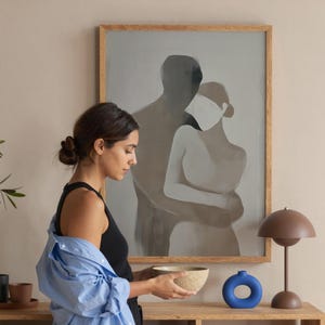 Könnte beinhalten: Eine Frau hält eine beige Schüssel in der Nähe eines Holzregals mit Büchern, Tassen und einem blauen Keramikring. Eine braune Lampe und ein gerahmtes Kunstwerk mit zwei Figuren befinden sich ebenfalls auf dem Regal. Die Frau trägt ein schwarzes Oberteil und ein hellblaues Hemd.