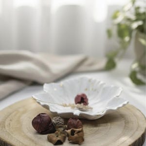 Könnte beinhalten: Eine weiße, blütenblattförmige Keramikschale mit einer kleinen, getrockneten Blume und dekorativen Elementen auf einer Holzscheibe. Die Anordnung umfasst getrocknete Knospen und Samen.