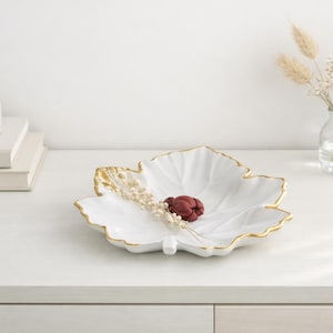 Könnte beinhalten: Eine weiße Keramikschale in Form eines Ahornblattes mit einem metallisch gold bemalten Rand. Die Schale weist geprägte Blattadern auf und enthält ein kleines Bündel getrockneter weißer Blumen sowie eine einzelne dunkelrote Blütenknospe.
