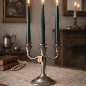Può includere: Un candelabro in metallo argentato a tre bracci che sostiene candele affusolate verde scuro. Ogni candela è accesa e produce una piccola fiamma. Una piccola etichetta rettangolare in carta beige è legata allo stelo centrale con un filo.