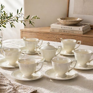 May include: A cream-coloured tea set comprising six cups, saucers, and a lidded sugar bowl. The cups and sugar bowl feature a ribbed design. The set is arranged on a white tablecloth, ideal for a formal tea setting.