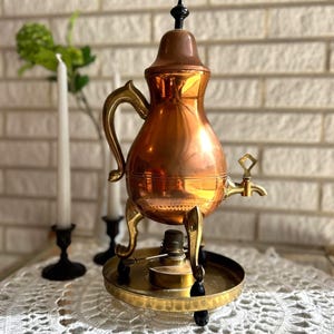 Op de afbeelding: Een vintage koperen en messing theepot met een zwarte knop. De ketel heeft een gebogen handvat en een kraan. Hij staat op een messing standaard met een brander en een ronde basis. De achtergrond omvat een witte bakstenen muur en kaarsen.