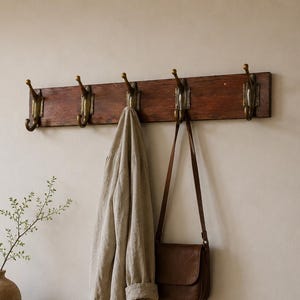 Op de afbeelding: Een rechthoekige wandkapstok van donker hout met vijf messingkleurige metalen haken. Een beige linnen overhemd en een bruine leren schoudertas hangen aan de haken. De kapstok is bevestigd aan een lichte muur.