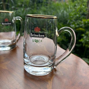 May include: Two clear glass beer mugs with gold-toned rims and handles. Each mug features the red Heineken logo, the word Heineken in black text on a white background, and a small green floral graphic printed on the front of the glass body.