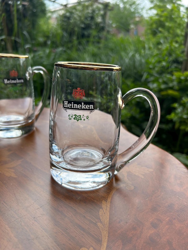 May include: Two clear glass beer mugs with gold-toned rims and handles. Each mug features the red Heineken logo, the word Heineken in black text on a white background, and a small green floral graphic printed on the front of the glass body.