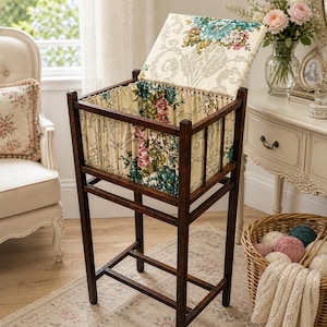 Puede incluir: Una cesta de costura de madera vintage con tapa con bisagras y forro de tela floral. El marco marrón oscuro sostiene una cesta rectangular con un interior de tela estampada. La tapa también está cubierta con un estampado floral.