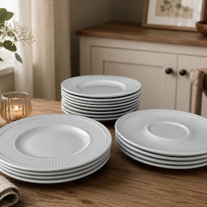 Op de afbeelding: Verschillende stapels witte keramische borden met een kralenrand en een iets verhoogd midden. De borden staan op een houten tafel, met een vaas met bloemen en een kandelaar op de achtergrond. De borden hebben een klassiek, elegant ontwerp.
