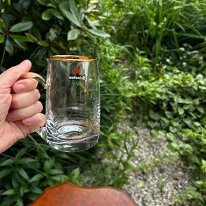 May include: A clear glass beer mug with a gold-rimmed top edge and a sturdy handle. The front of the glass features the red Heineken star logo and the brand name printed in white text. The glass has a thick, weighted base and smooth sides.