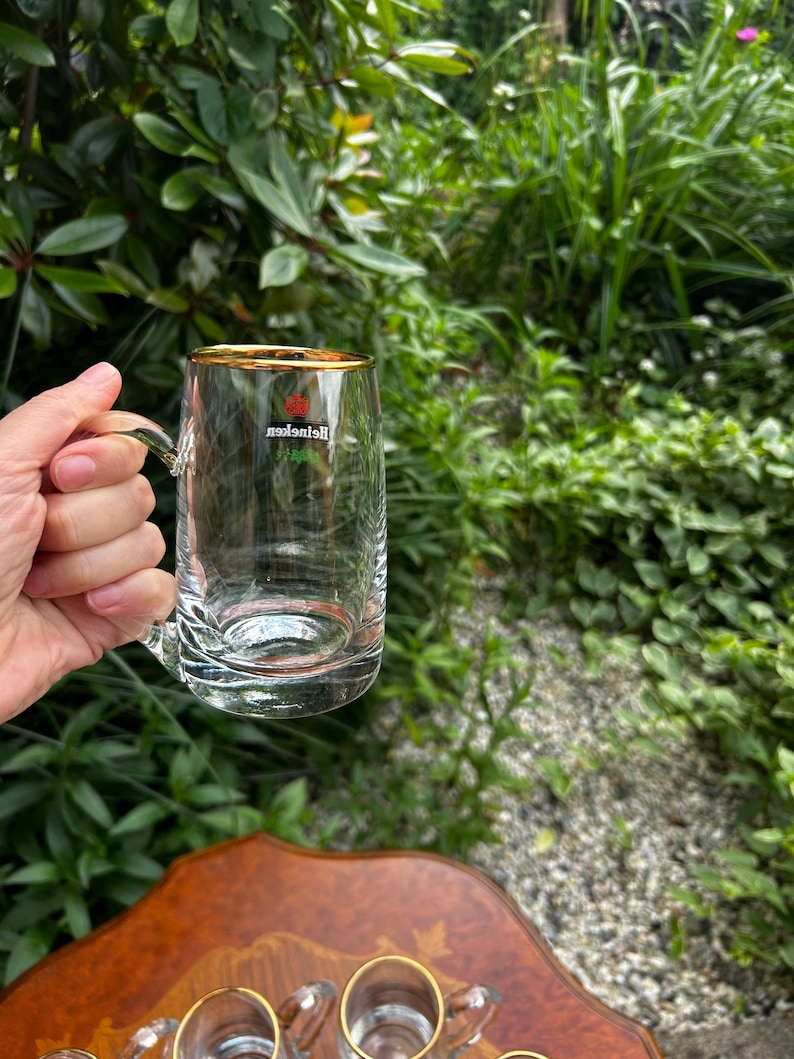 May include: A clear glass beer mug with a gold-rimmed top edge and a sturdy handle. The front of the glass features the red Heineken star logo and the brand name printed in white text. The glass has a thick, weighted base and smooth sides.