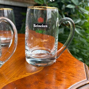 May include: A clear glass beer mug with a sturdy handle and a gold-colored rim. The front of the mug features the red Heineken logo, the word Heineken in black text, and a small green decorative graphic element printed on the glass surface.