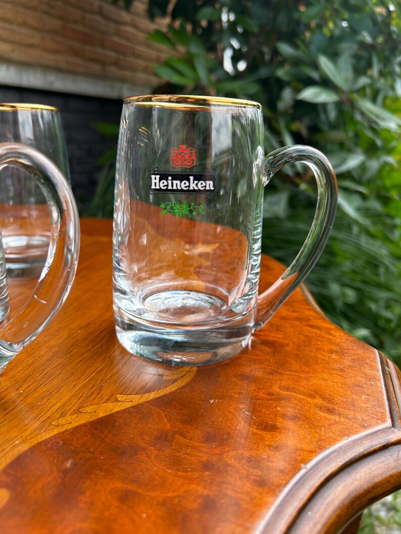 May include: A clear glass beer mug with a sturdy handle and a gold-colored rim. The front of the mug features the red Heineken logo, the word Heineken in black text, and a small green decorative graphic element printed on the glass surface.