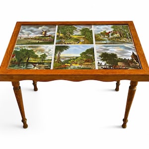 Op de afbeelding: Een vintage houten bijzettafel met zes landschapstegels. De tafel heeft een rechthoekig blad en vier gedraaide poten. De tegels tonen scènes met windmolens, huizen en waterwegen. De tafel is ongeveer 60 cm lang.