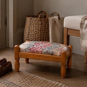Op de afbeelding: Een houten voetenbankje met een bloemenpatroon kussen. Het frame is lichtbruin en heeft vier afgeronde poten. Een paar bruine leren loafers staat op een vloerkleed voor de voetenbank. Een gevlochten mand en een stoel staan op de achtergrond.
