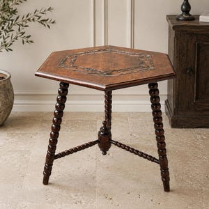 Op de afbeelding: Een zeshoekige houten bijzettafel met een donkerbruine afwerking. Het tafelblad heeft een centraal paneel met houtnerf omringd door een ingelegde rand. De tafel staat op drie poten met een gedraaid, kraalachtig ontwerp en een centrale steun.