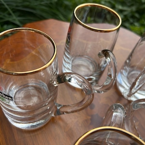 May include: A set of clear glass beer mugs featuring a gold rim at the top and a curved handle. Each mug has horizontal ridges along the lower body and displays the Heineken logo in red, green, and white on the side of the glass surface.