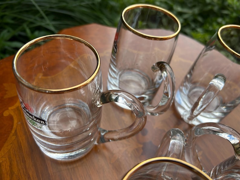 May include: A set of clear glass beer mugs featuring a gold rim at the top and a curved handle. Each mug has horizontal ridges along the lower body and displays the Heineken logo in red, green, and white on the side of the glass surface.