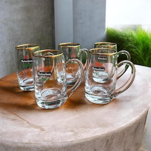 May include: A set of four clear glass beer mugs with handles, each featuring a gold-rimmed top edge and the Heineken logo printed in black, red, and green on the front. The mugs are arranged in a cluster, showcasing their transparent glass construction.