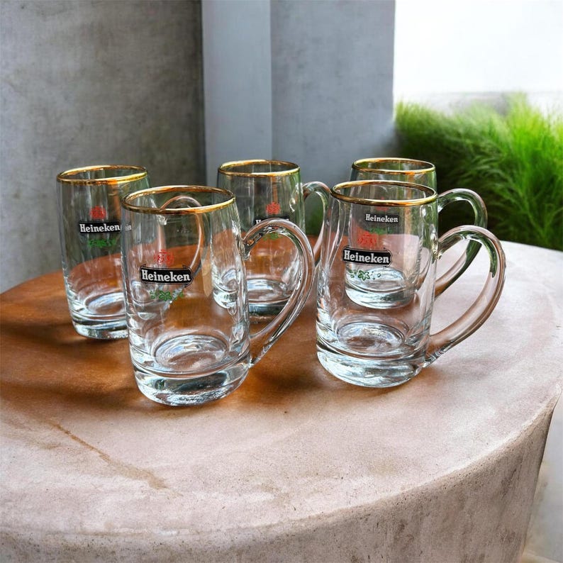 May include: A set of four clear glass beer mugs with handles, each featuring a gold-rimmed top edge and the Heineken logo printed in black, red, and green on the front. The mugs are arranged in a cluster, showcasing their transparent glass construction.