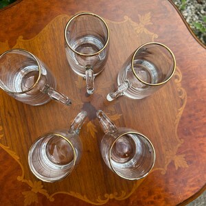 May include: A set of five clear glass mugs arranged in a circular pattern. Each mug features a cylindrical body with horizontal ribbed detailing and a smooth glass handle. The top rim of every mug is finished with a thin, metallic gold band.