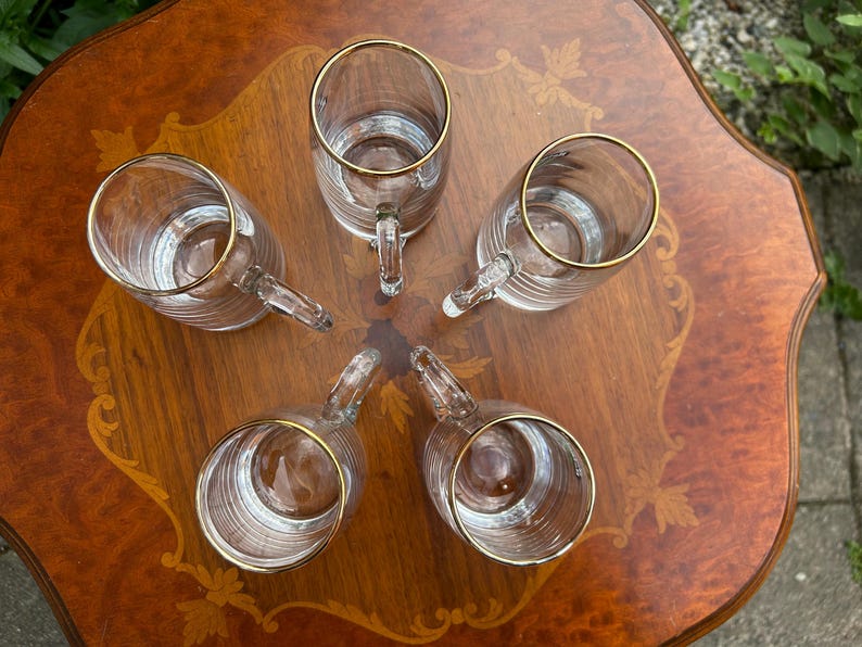 May include: A set of five clear glass mugs arranged in a circular pattern. Each mug features a cylindrical body with horizontal ribbed detailing and a smooth glass handle. The top rim of every mug is finished with a thin, metallic gold band.