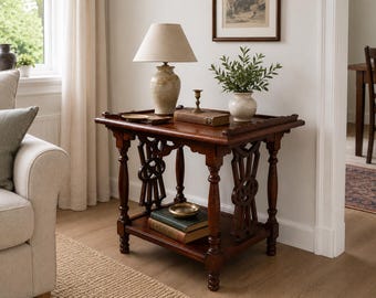 Peut inclure: Une table d'appoint rectangulaire en bois foncé avec une étagère inférieure, des pieds tournés et des panneaux latéraux décoratifs. Le plateau supporte une lampe beige, un petit bougeoir en laiton et un livre. L'étagère contient deux livres.