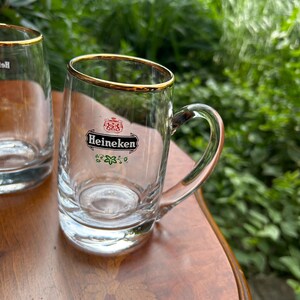 May include: A clear glass beer mug featuring a gold-trimmed rim and a sturdy handle. The front of the glass displays the Heineken logo in black, red, and green, including the brand name and a small crest above it with a floral design below.