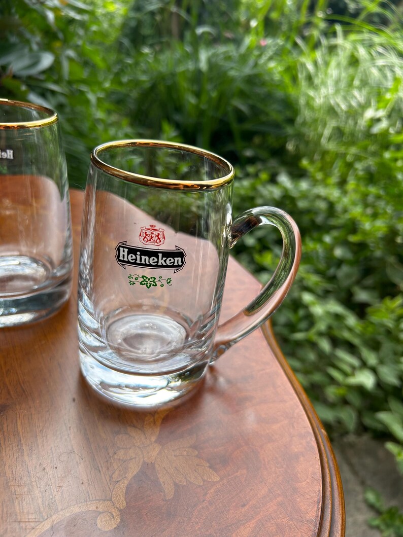 May include: A clear glass beer mug featuring a gold-trimmed rim and a sturdy handle. The front of the glass displays the Heineken logo in black, red, and green, including the brand name and a small crest above it with a floral design below.