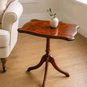 Puede incluir: Una mesa auxiliar de pedestal de madera con una superficie rectangular de chapa de madera de raíz marrón oscuro y bordes festoneados. La mesa se apoya en una columna central torneada con tres patas curvas. Un pequeño jarrón de cerámica color crema descansa sobre ella.
