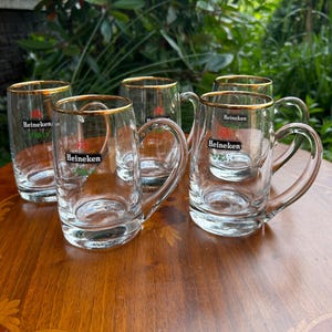 May include: A set of four clear glass beer mugs with gold-toned rims. Each mug features a handle and the Heineken logo printed in black, red, and green on the front. The glass mugs are arranged in a group, showcasing their transparent design.