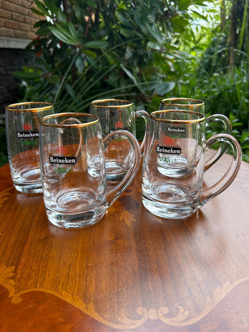 May include: A set of four clear glass beer mugs with gold-toned rims. Each mug features a handle and the Heineken logo printed in black, red, and green on the front. The glass mugs are arranged in a group, showcasing their transparent design.