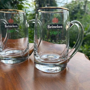 May include: A set of clear glass beer mugs with handles, each featuring a gold rim and the Heineken logo printed in black, red, and green on the front. The mugs have a thick, sturdy base and a smooth, cylindrical body design for serving beverages.