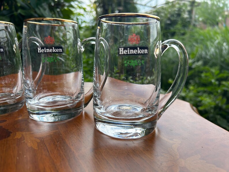 May include: A set of clear glass beer mugs with handles, each featuring a gold rim and the Heineken logo printed in black, red, and green on the front. The mugs have a thick, sturdy base and a smooth, cylindrical body design for serving beverages.