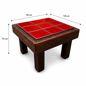 Op de afbeelding: Een kleine, vierkante houten kruk met een rood betegeld blad. De kruk is 18 cm breed, 18 cm lang en 14 cm hoog. Het donkerbruine houten frame contrasteert met de helderrode tegels.