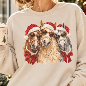 Puede incluir: Una sudadera beige de cuello redondo con un gráfico de tres caballos con gorros de Papá Noel, gafas de sol con estampado de leopardo y collares de perlas con lazos rojos, acompañada de una taza blanca con árboles de Navidad.