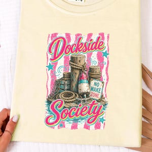 May include: A pale yellow t-shirt featuring a graphic print of wooden dock pilings, a buoy, rope, and a sign that reads NO WAKE ZONE. The text Dockside Society is printed in pink and blue script above and below the nautical illustration.