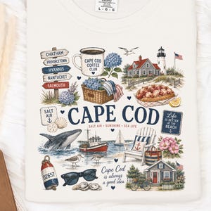Puede incluir: Una camiseta de color crema con un estampado gráfico de imágenes costeras de Cape Cod. El diseño incluye un faro, una ballena, un rollo de langosta, una silla de playa y señales que apuntan a pueblos como Chatham y Provincetown con el texto Cape Cod.