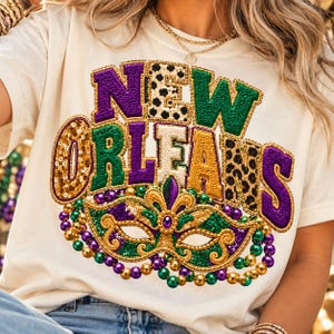 Puede incluir: Una camiseta color crema con el texto NEW ORLEANS en letras grandes, texturizadas y multicolores. El diseño incluye una máscara de lentejuelas, cuentas doradas, verdes y moradas, y patrones de leopardo y lunares.