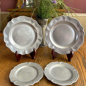 May include: A set of four silver-toned metal plates with a matte finish and scalloped edges. Two larger plates are displayed upright on dark wooden stands, while two smaller plates are positioned flat in front of them on a wooden surface.