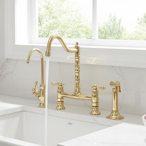 May include: A gold-coloured kitchen tap with a bridge design, featuring a gooseneck spout and separate sprayer. The tap is installed over a white farmhouse sink. A white marble countertop and a window are in the background.